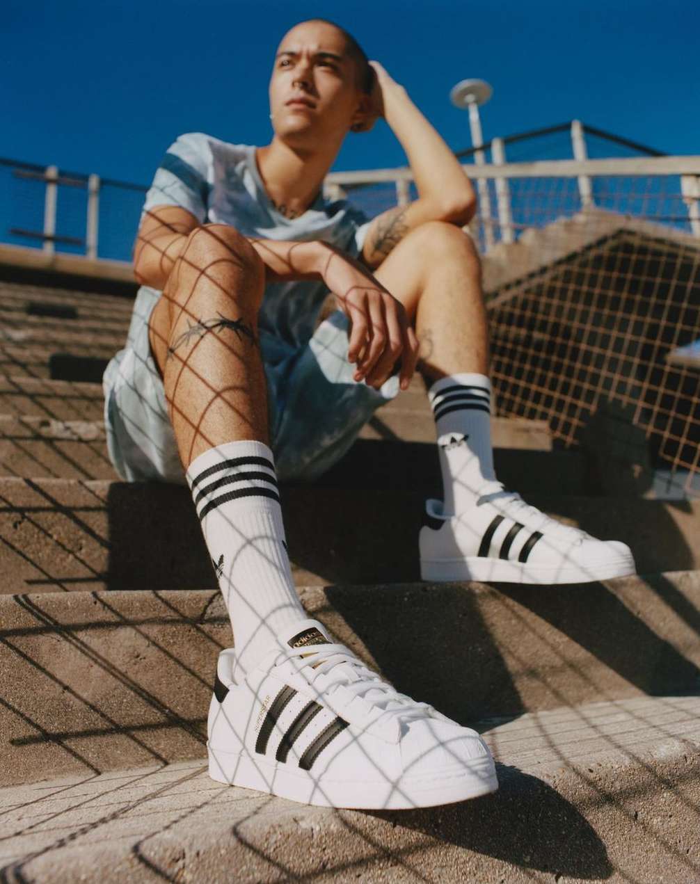 Young adult sits on concrete steps, wearing a light blue shirt, shorts, black and white socks, and Adidas sneakers on a sunny day.