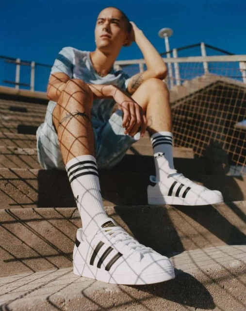 Young adult sits on concrete steps, wearing a light blue shirt, shorts, black and white socks, and Adidas sneakers on a sunny day.