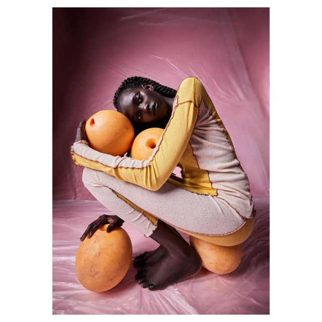 Woman with dark skin and long braids poses crouching with decorative orange spheres against a pink studio backdrop wearing knitwear.