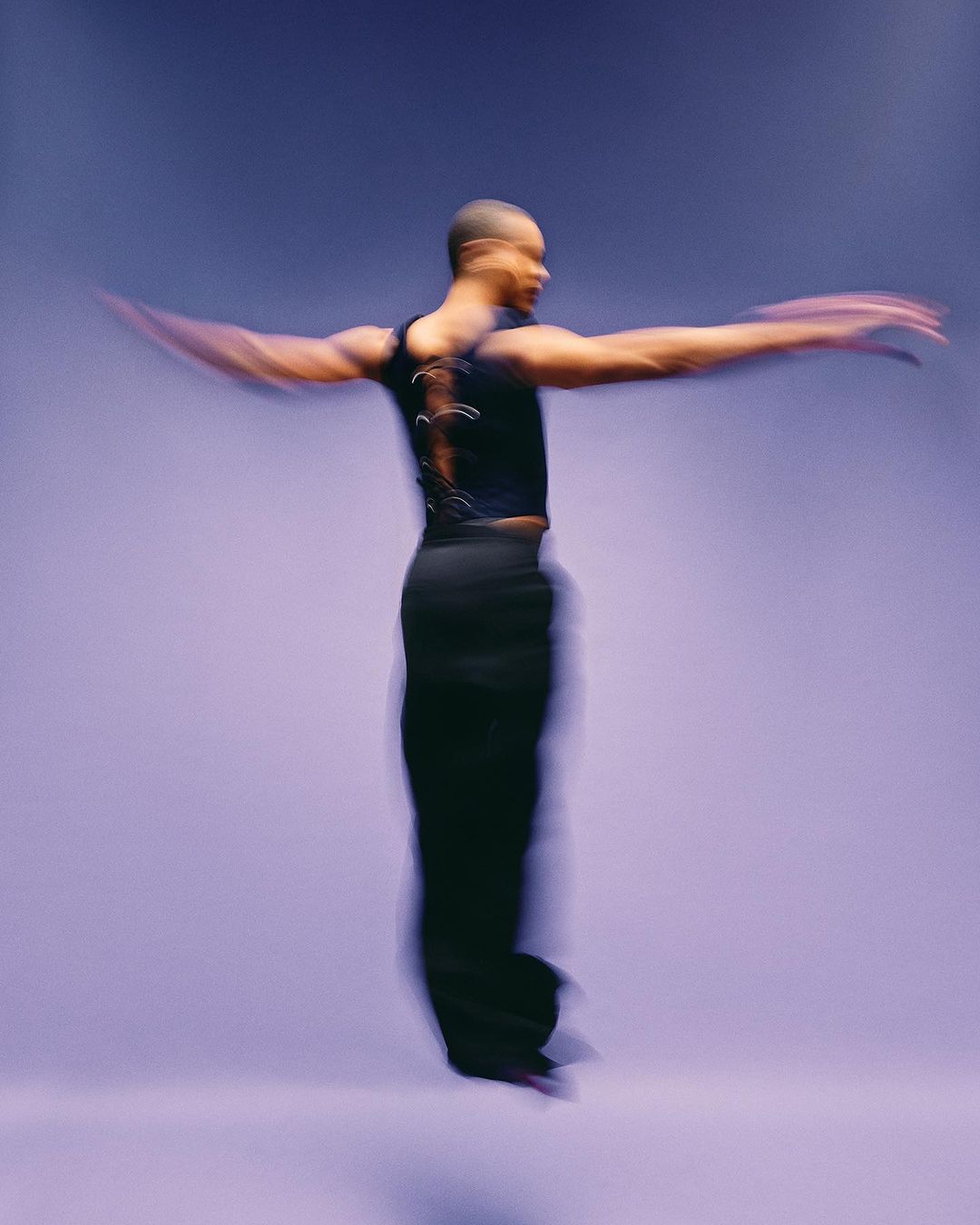 Adult ballet dancer in mid-air wearing black top and trousers performing a dance routine against a light purple gradient background.