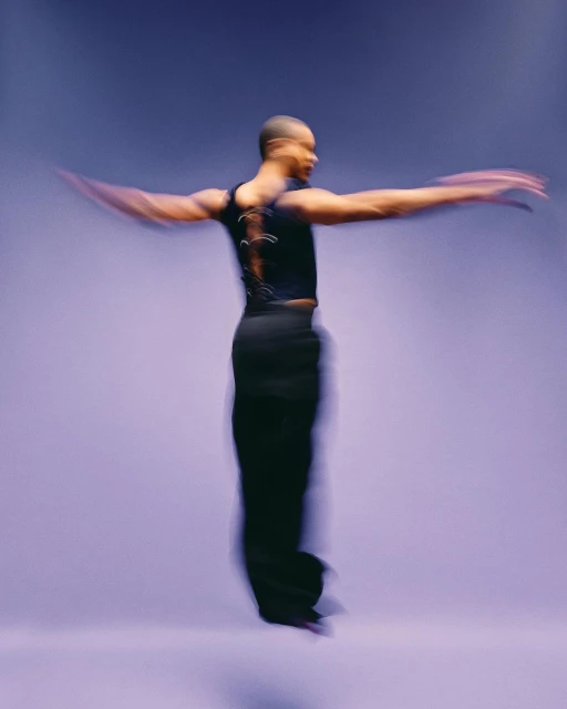 Adult ballet dancer in mid-air wearing black top and trousers performing a dance routine against a light purple gradient background.