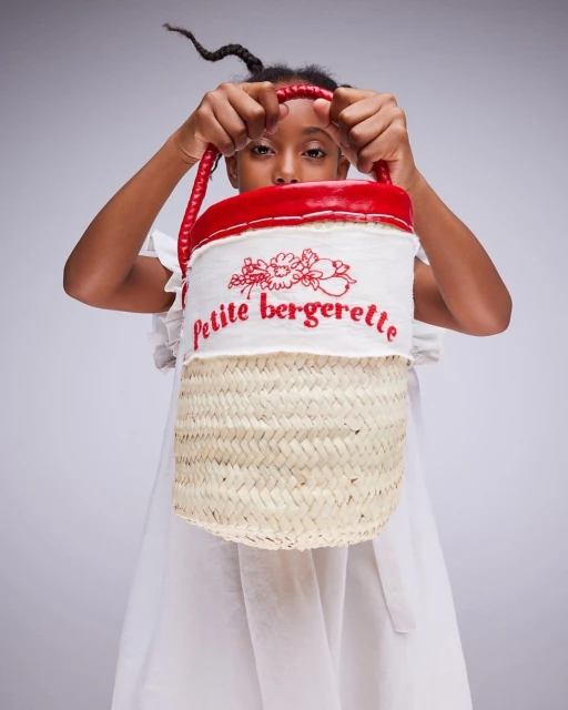 Young girl poses in a photo studio, showcasing a petite bergreette straw bag with red trim and handle. The background is a light gray.