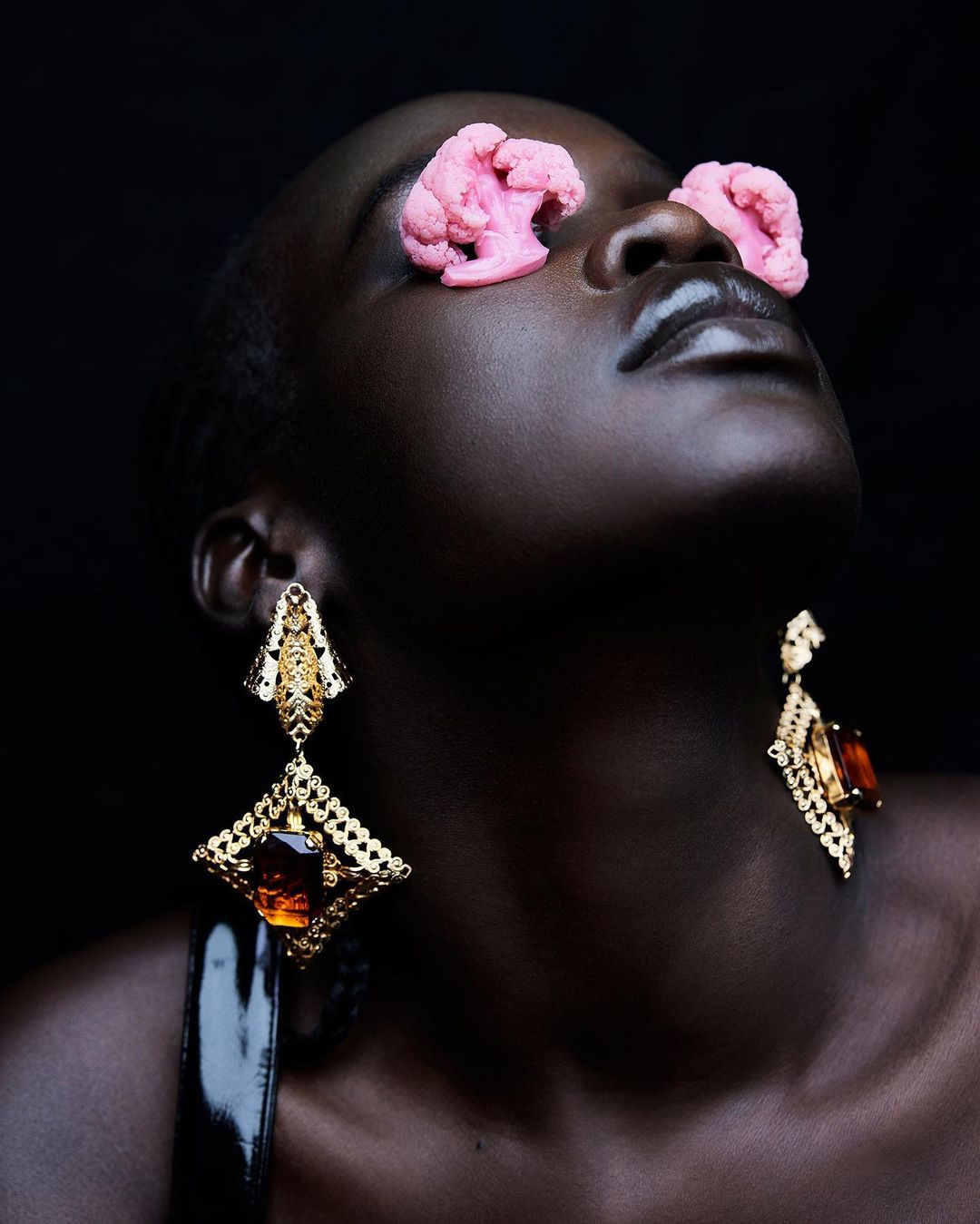 Close-up studio shot of a stylish Black woman wearing large gold earrings and pink cauliflower on her eyes against a dark background.