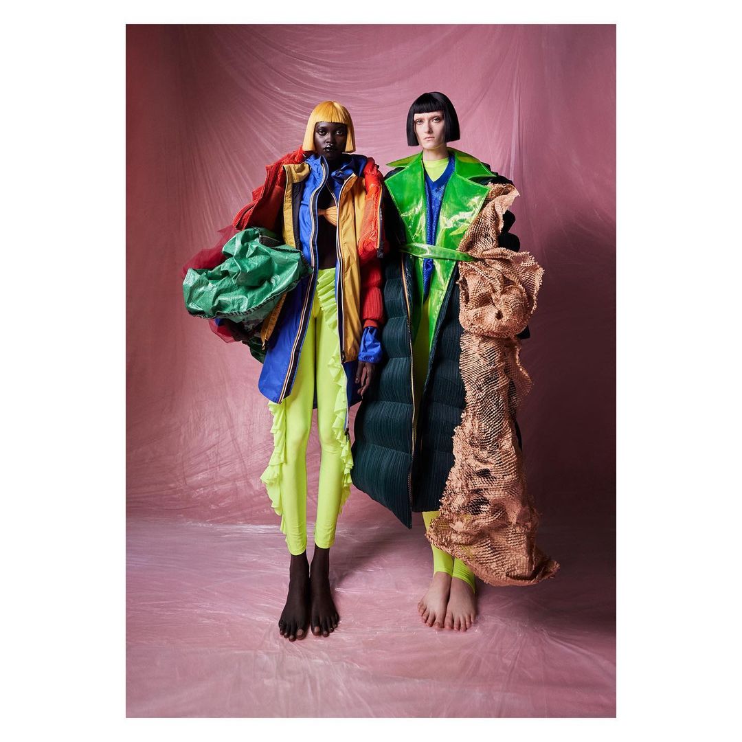 Two fashion models pose in colorful, unconventional clothing against a pink backdrop, showcasing unique and eye-catching designs.