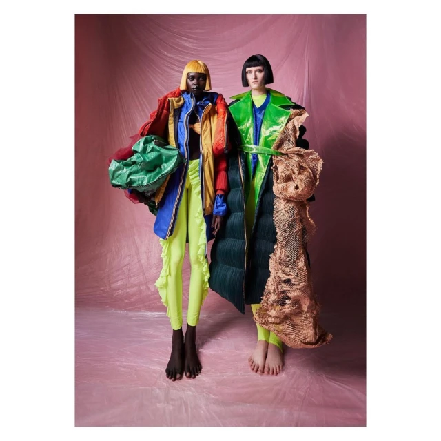 Two fashion models pose in colorful, unconventional clothing against a pink backdrop, showcasing unique and eye-catching designs.