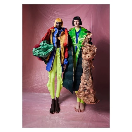 Two fashion models pose in colorful, unconventional clothing against a pink backdrop, showcasing unique and eye-catching designs.