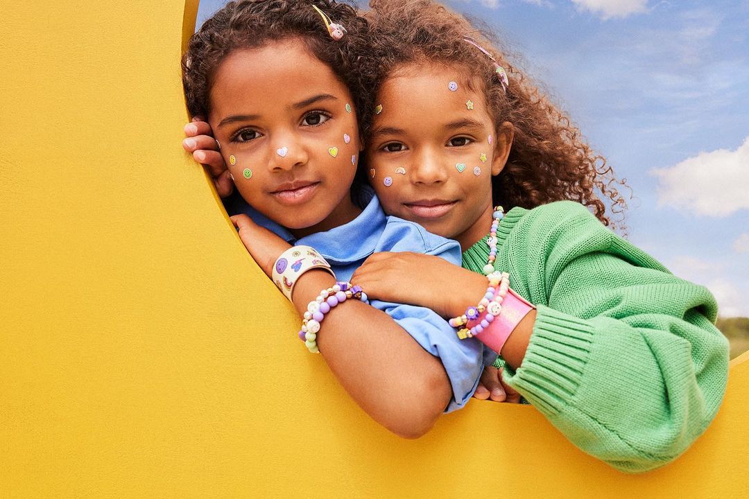 Two young girls with stickers on their faces pose together in front of a yellow structure and a partly cloudy sky.