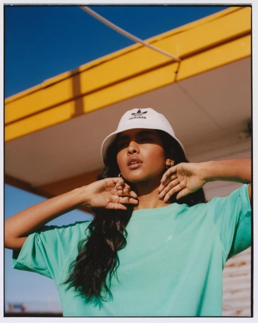 An Indian woman models an Adidas bucket hat and green tee in a sunlit outdoor setting with a blue sky and yellow building.
