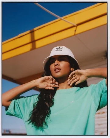 An Indian woman models an Adidas bucket hat and green tee in a sunlit outdoor setting with a blue sky and yellow building.