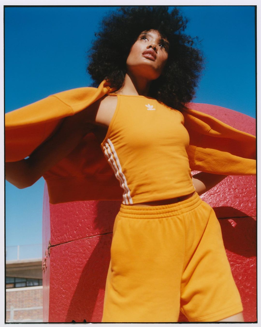 Woman with afro hairstyle wears an orange Adidas athletic set posing outdoors against a vibrant blue sky and red geometric structure.
