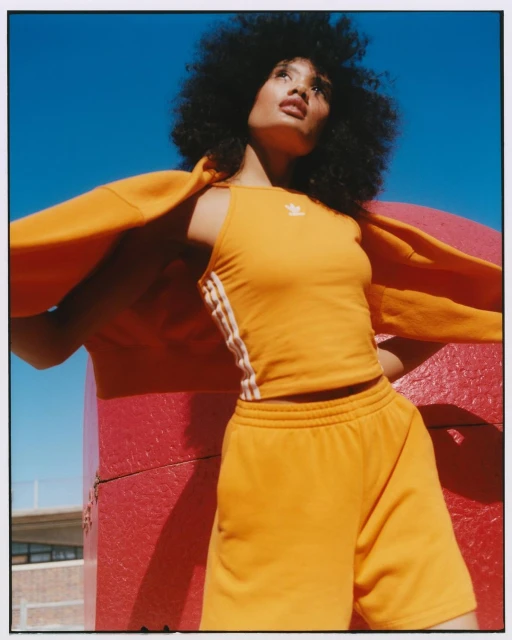 Woman with afro hairstyle wears an orange Adidas athletic set posing outdoors against a vibrant blue sky and red geometric structure.