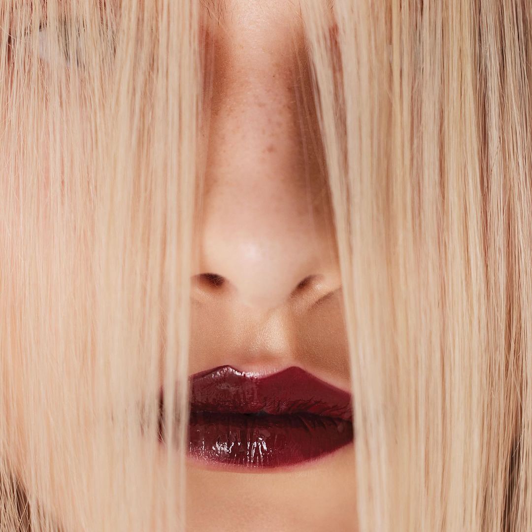 Close-up of a woman's face with blonde hair partially covering her face and shiny, dark red lipstick on her lips.