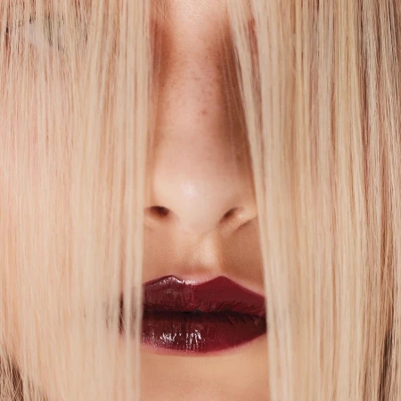 Close-up of a woman's face with blonde hair partially covering her face and shiny, dark red lipstick on her lips.
