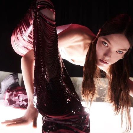 An adult female with long brown hair and pale skin poses with futuristic fashion, against a dark background, using an eye-level angle.