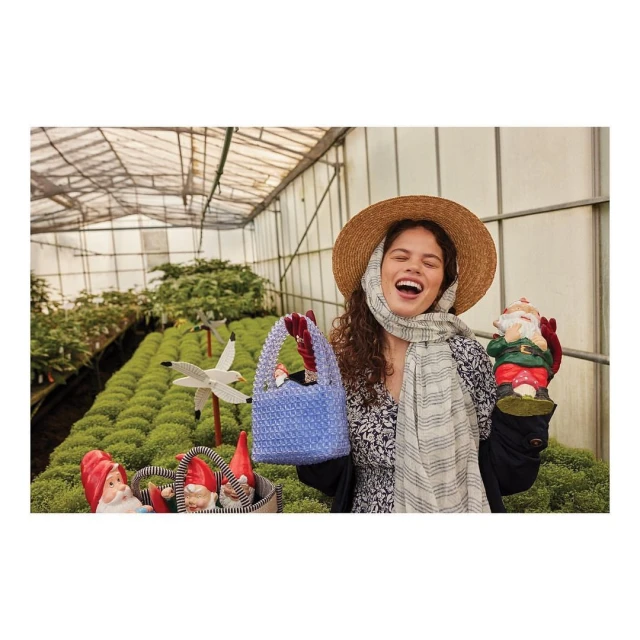 Woman in straw hat laughs while holding a blue beaded bag and a gnome figurine in greenhouse, surrounded by rows of green plants.