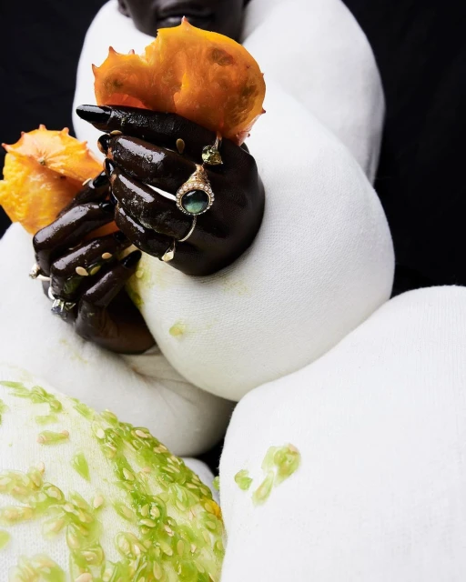 Close-up studio shot features a person with dark skin wearing jewelry and holding horned melon against a textured white surface.
