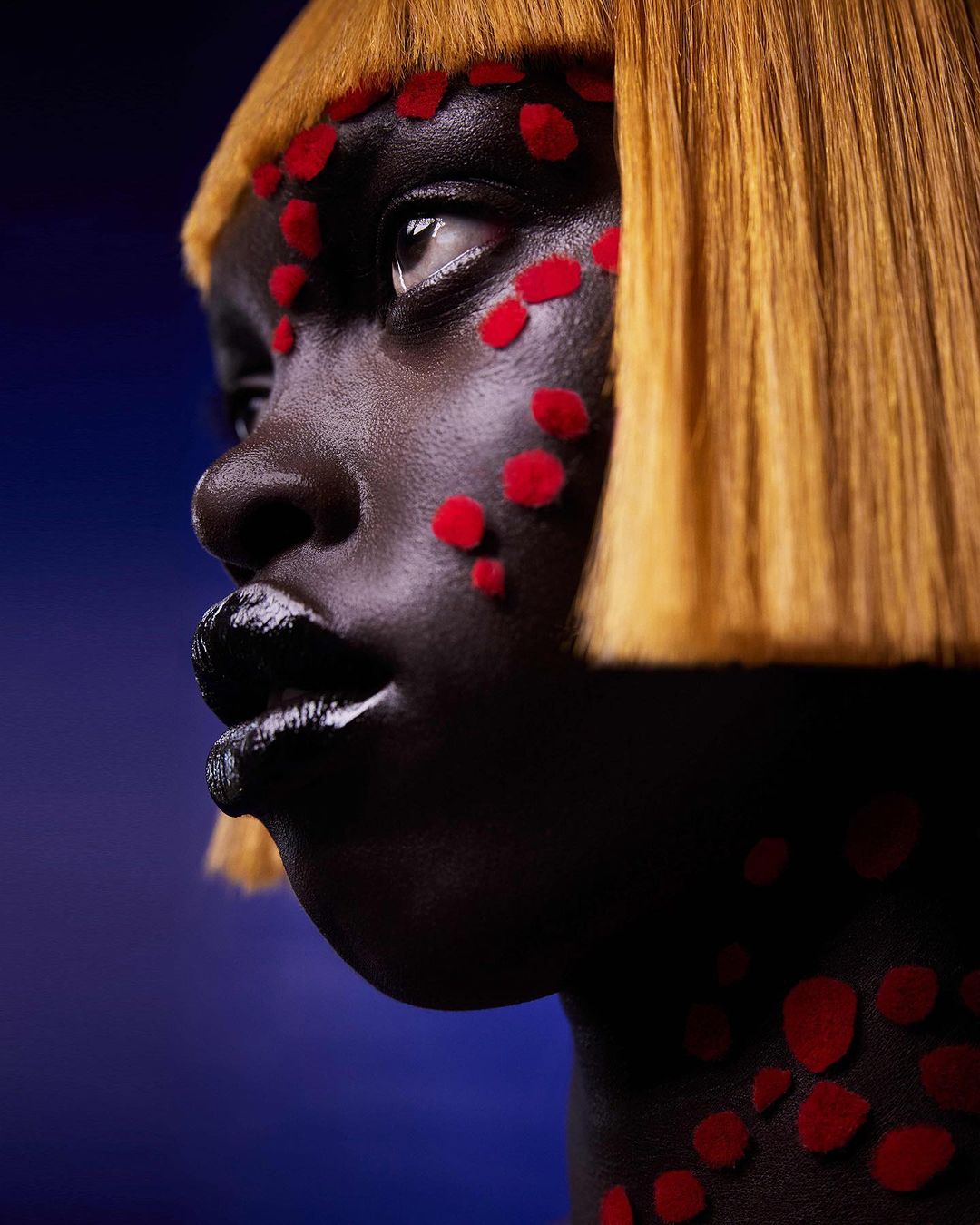 Close-up of a striking African female model with dark skin, a blonde bob, and red geometric face paint against a blue gradient backdrop.