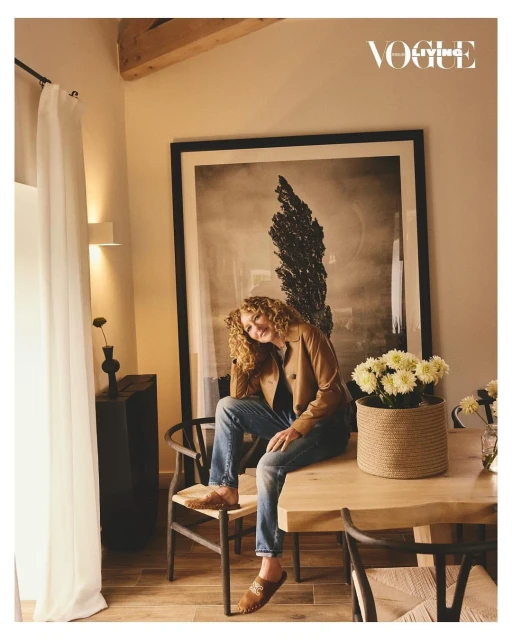 Adult female seated on a wood table in a room with a framed photo, flowers, wood chairs, and a window with white curtains.