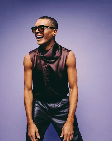 Person smiles wearing sunglasses and a sleeveless top in a studio shot against a solid purple background. Fashion and portrait photography.