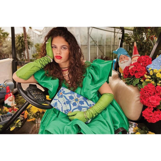 Brunette woman in a green dress, elbow on the steering wheel of a golf cart, holding a clutch, with garden gnomes in the background.