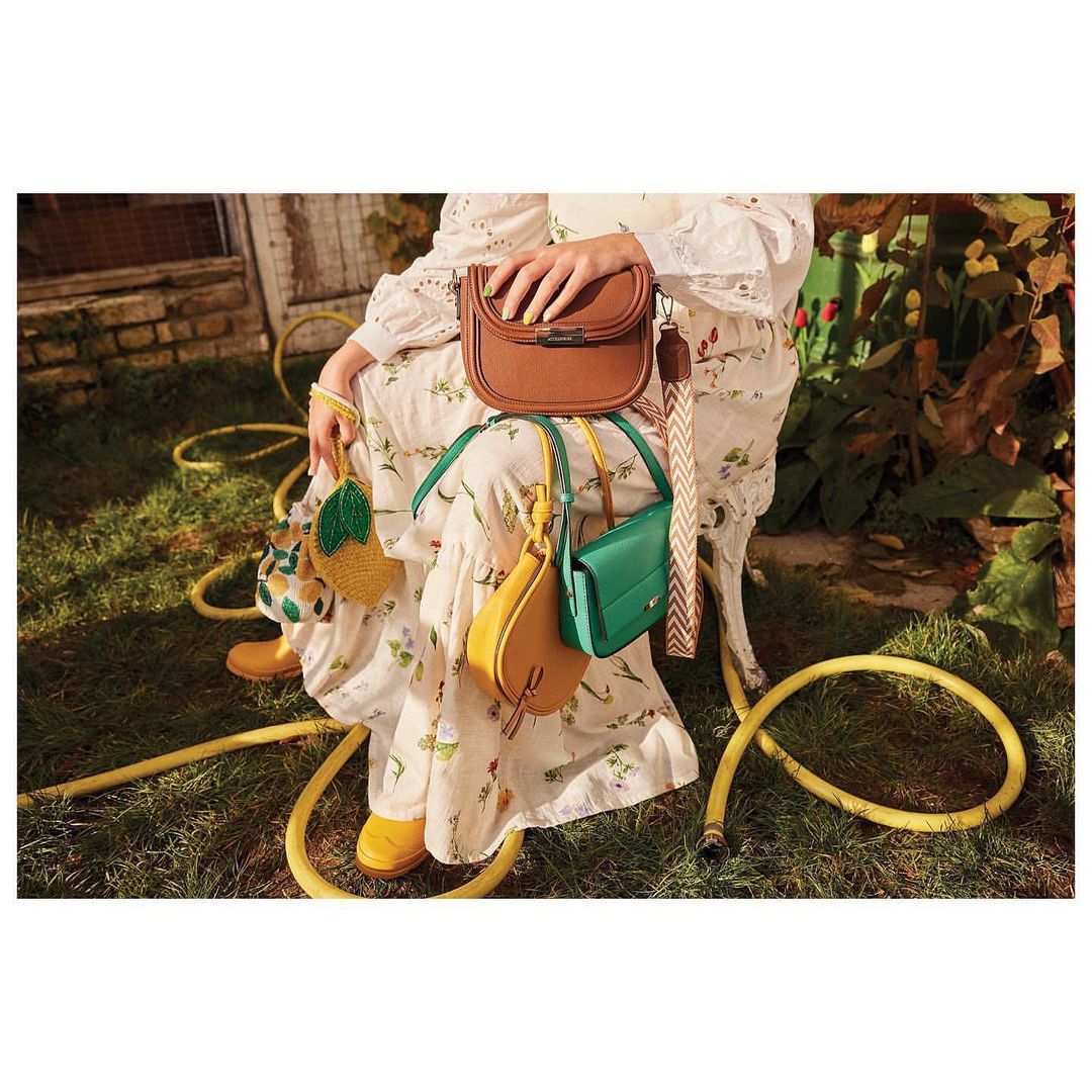 Woman seated outdoors wearing a floral dress, holding brown handbag, with yellow and green handbags and yellow rubber boots.