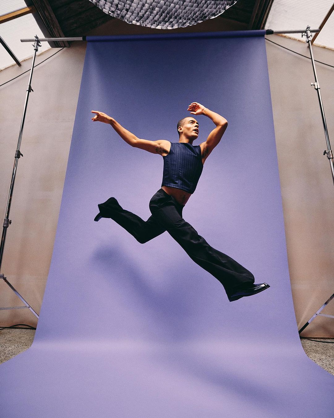Man in mid-air jump wearing a striped vest and black pants against a studio backdrop, showcasing dynamic movement and fashion.