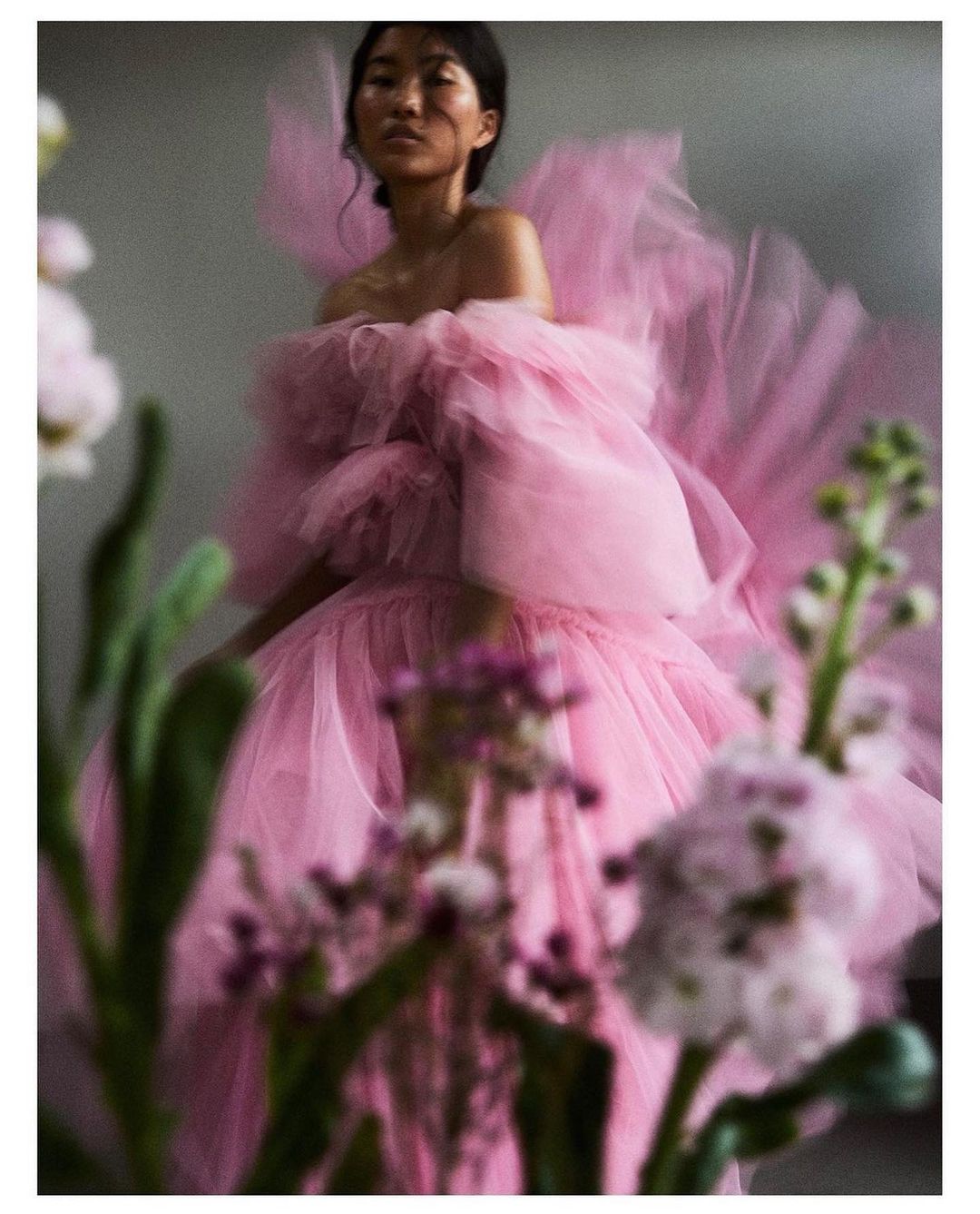A woman poses in a pink tulle dress with plants in the foreground. The photo features soft lighting and a muted color palette.