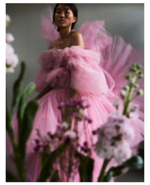 A woman poses in a pink tulle dress with plants in the foreground. The photo features soft lighting and a muted color palette.
