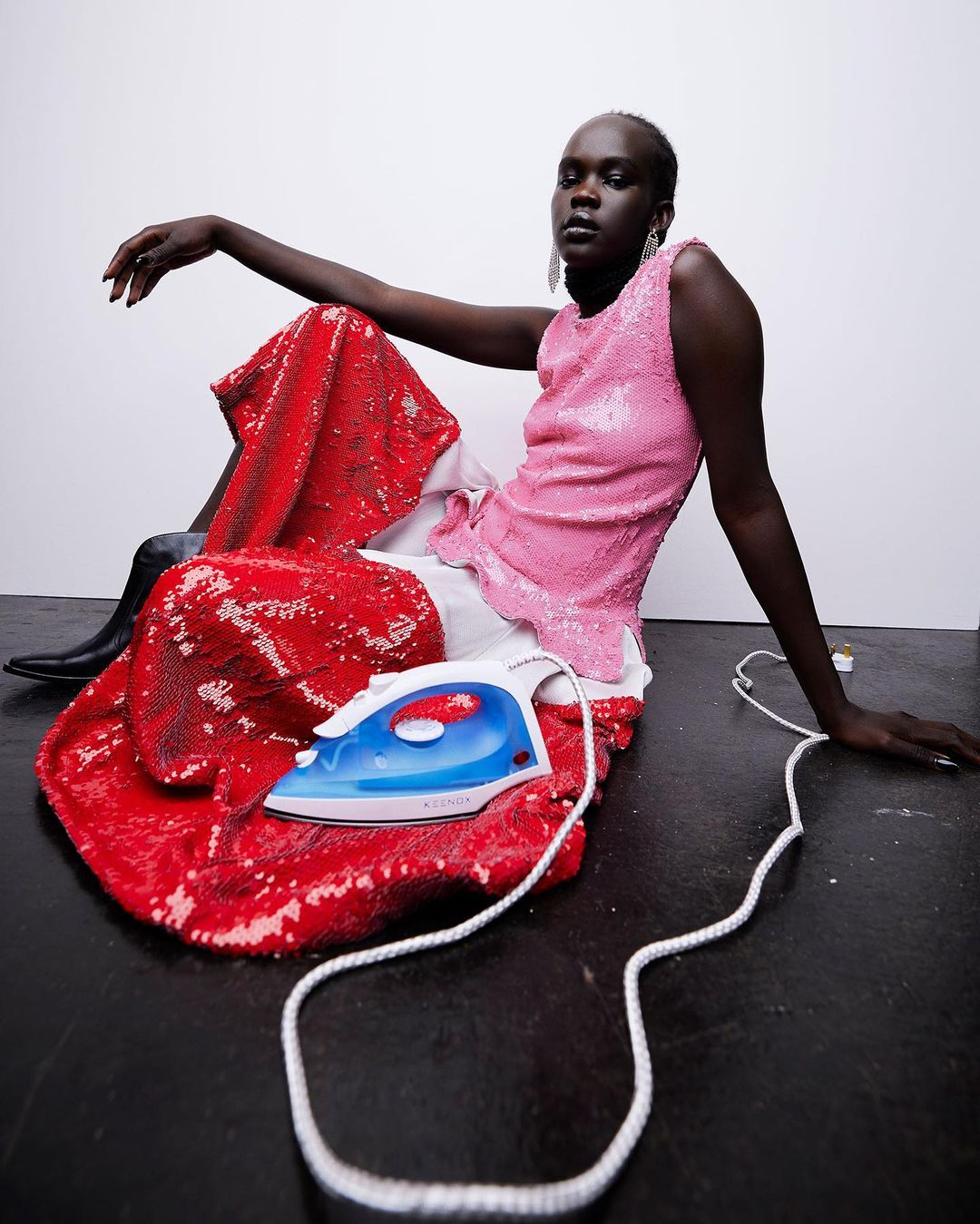 Fashionable adult female poses on the floor with a clothing iron, wearing a sequined pink top and red pants in a studio.