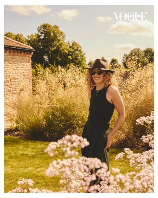 Woman in a hat and black outfit stands in a lush garden with a brick building visible. Vogue logo is placed in the upper right corner.