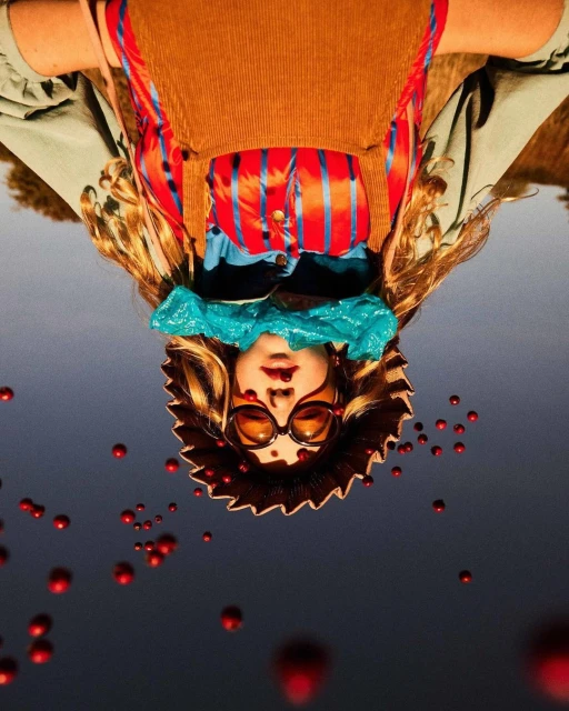 Upside-down portrait of a person wearing sunglasses, a straw hat, and a colorful outfit, surrounded by floating red orbs in a gray sky.
