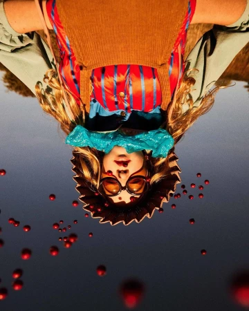 Upside-down portrait of a person wearing sunglasses, a straw hat, and a colorful outfit, surrounded by floating red orbs in a gray sky.