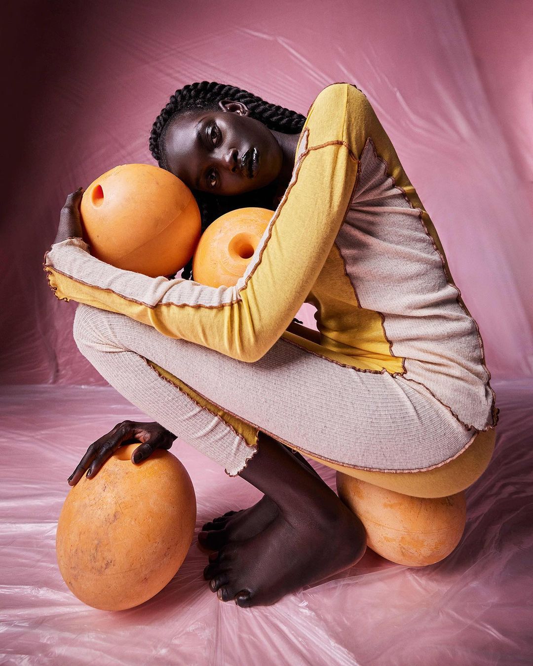 A dark-skinned woman in yellow and cream clothing squats surrounded by orange exercise balls against a pink, plastic-covered background.