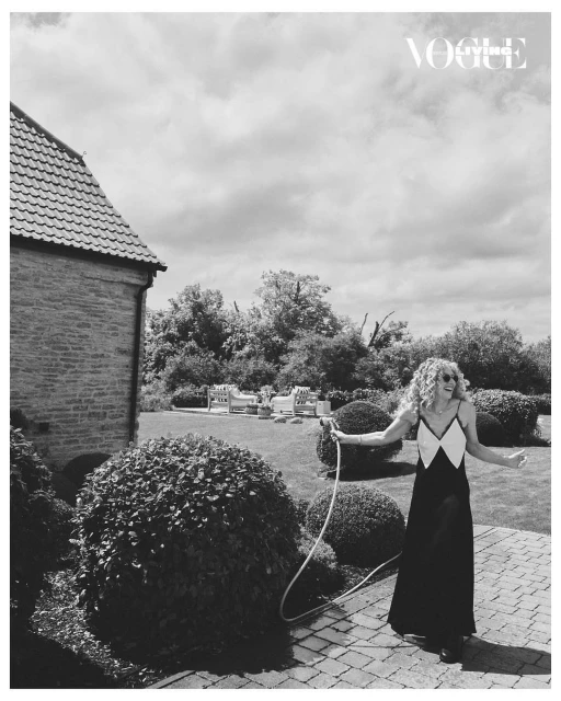 Woman in a long dress poses on a stone patio holding a garden hose with a brick house and manicured lawn in the background.