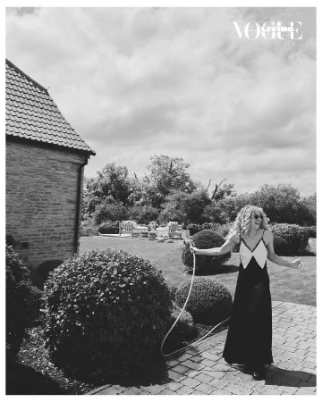 Woman in a long dress poses on a stone patio holding a garden hose with a brick house and manicured lawn in the background.