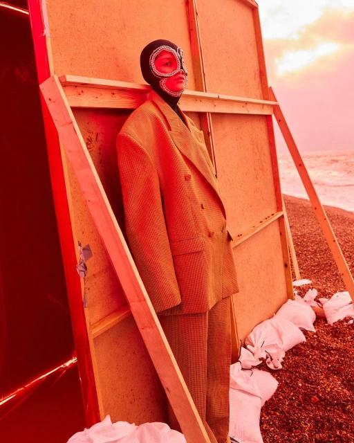 Person in suit and beaded mask stands next to wooden structure on pebble beach with sky in the background in a red color grade.