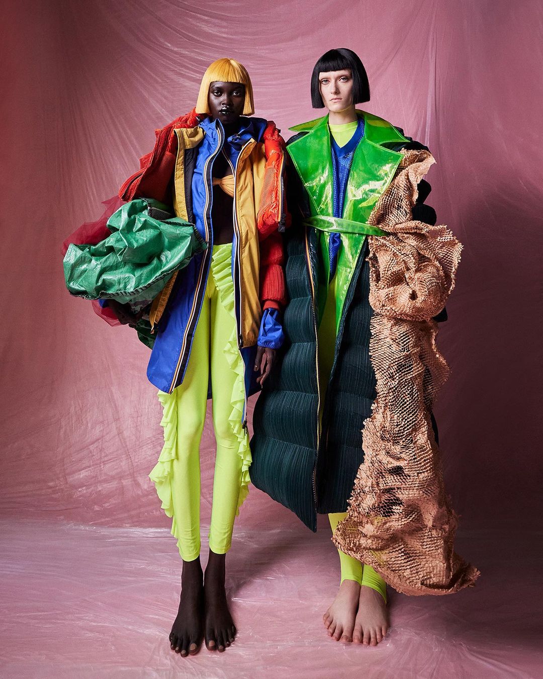 Two fashion models in unique, colorful outerwear, set against a pink backdrop, showcase avant-garde style and design. Striking presentation.