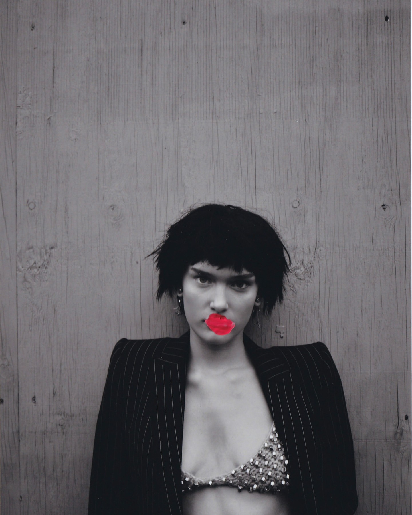 Adult female with short dark hair and red lips posing against a gray wooden background wearing a pinstripe suit jacket and a beaded bra top.