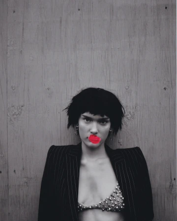 Adult female with short dark hair and red lips posing against a gray wooden background wearing a pinstripe suit jacket and a beaded bra top.
