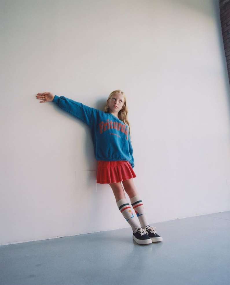 A young, blonde girl leans against a white wall, wearing a blue sweatshirt, red skirt, striped socks, and black platform shoes.