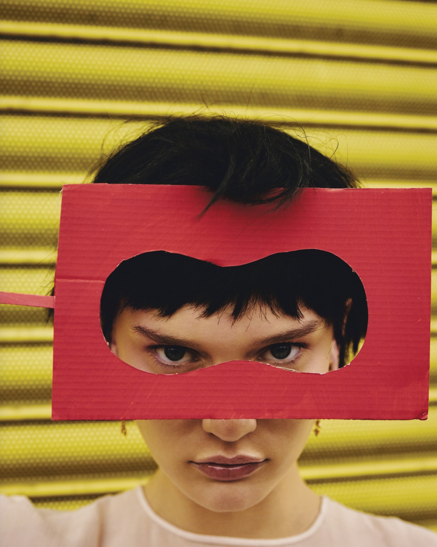 Close-up of a young woman with short dark hair holding a red cardboard mask with a heart-shaped eye cutout over her eyes indoors.