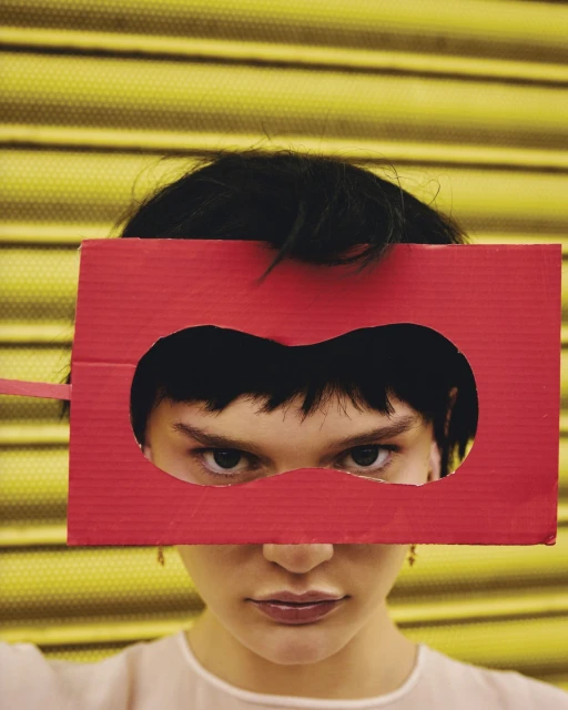 Close-up of a young woman with short dark hair holding a red cardboard mask with a heart-shaped eye cutout over her eyes indoors.