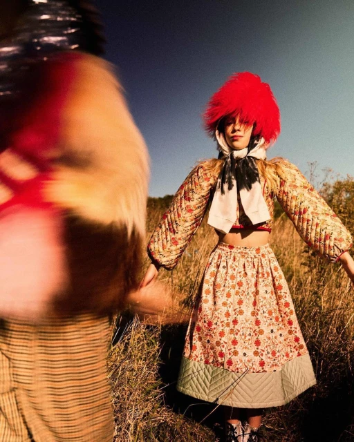 Two people in a field, one with a bright red fur hat and patterned dress, standing in a field of golden grasses under a clear blue sky.