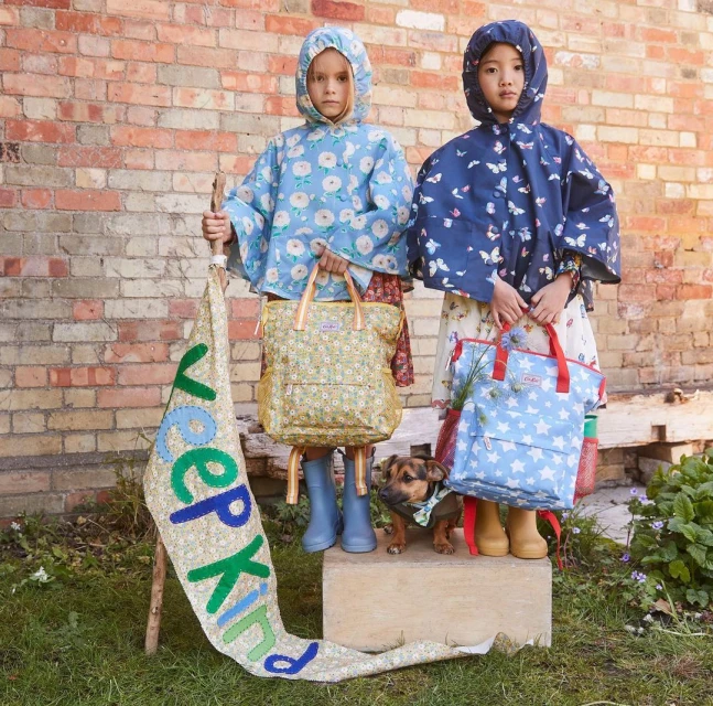 Two children in patterned rain ponchos and boots stand with bags and a dog in front of a brick wall, holding a sign.