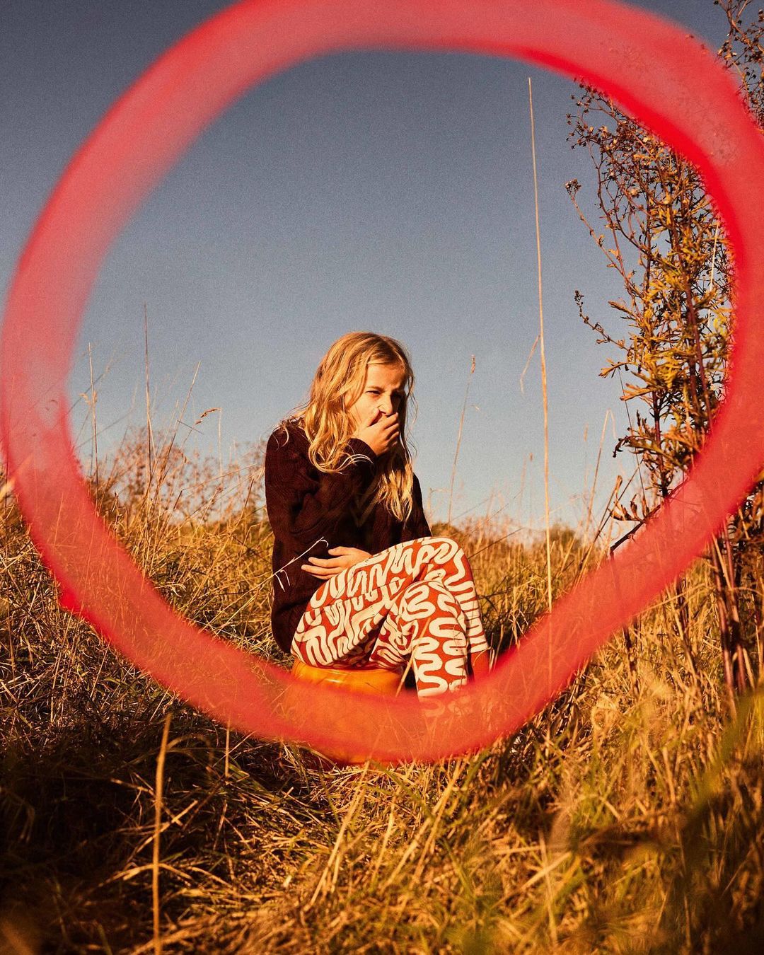 A young woman sits encircled by a red, out-of-focus ring in a grassy field, wearing patterned leggings and a dark sweater, enjoying the outdoors.