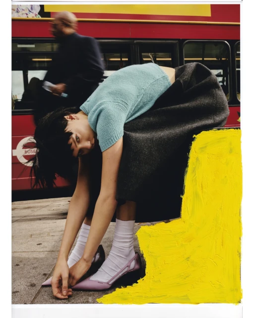 Fashion editorial shot: woman in stylish skirt and top bending over near red bus in London, featuring bold yellow painted accent.