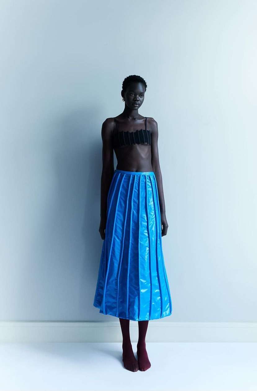 A fashion shot of a slender, dark-skinned adult woman wearing a black crop top and shiny blue skirt, posing against a muted blue wall.