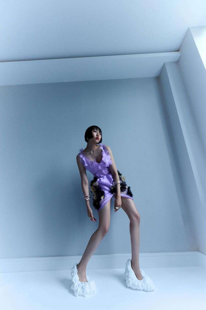 Woman poses in a studio wearing a lilac-colored ruffle dress and white ruffle shoes against a light blue wall and white baseboard.