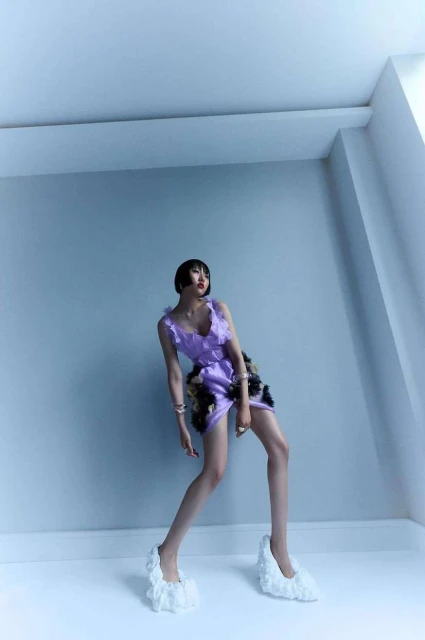 Woman poses in a studio wearing a lilac-colored ruffle dress and white ruffle shoes against a light blue wall and white baseboard.