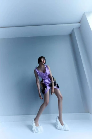 Woman poses in a studio wearing a lilac-colored ruffle dress and white ruffle shoes against a light blue wall and white baseboard.